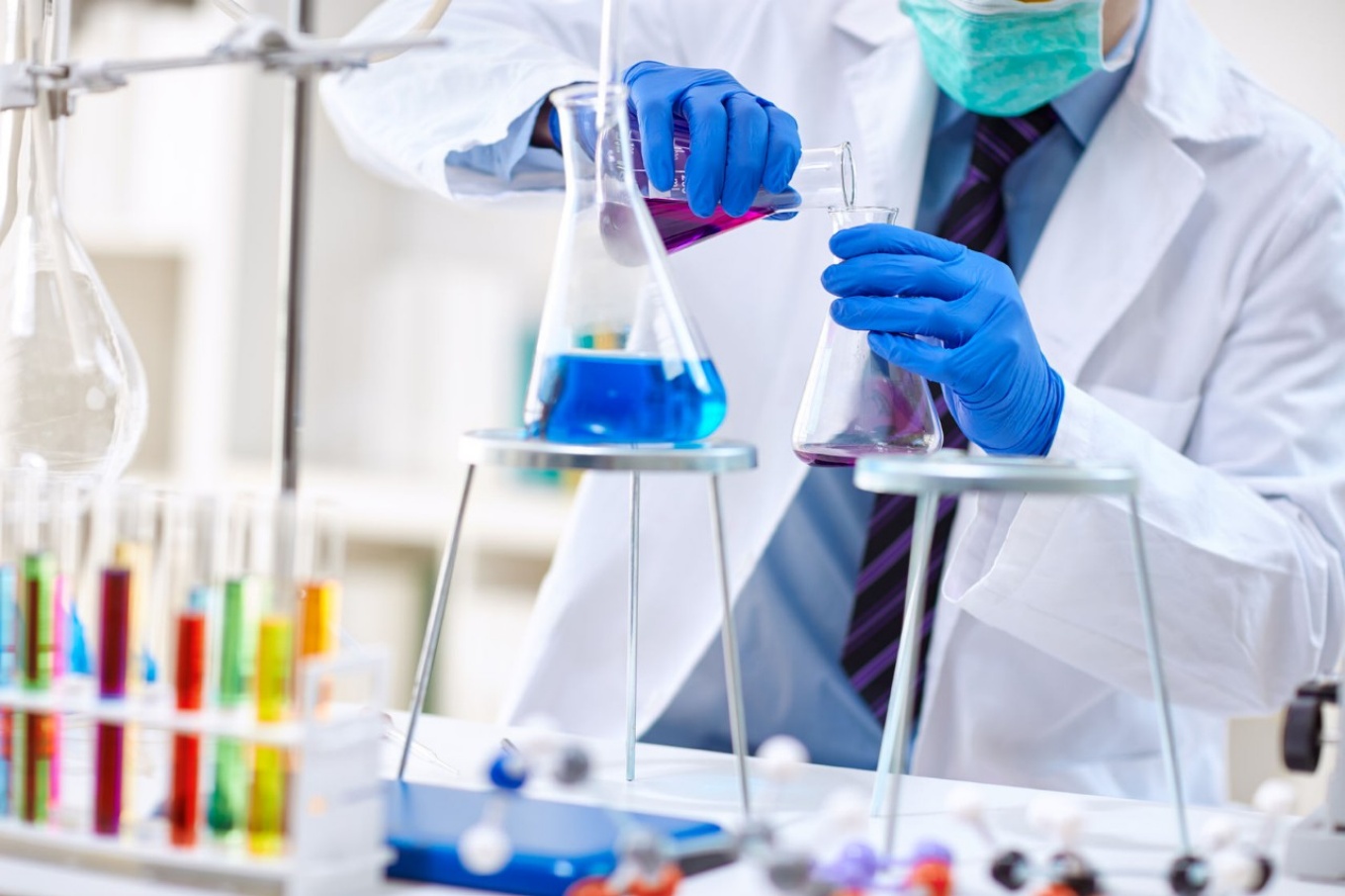 Person wearing a lab coat, shirt, tie and face mask, mixing colorful chemicals in a lab. 