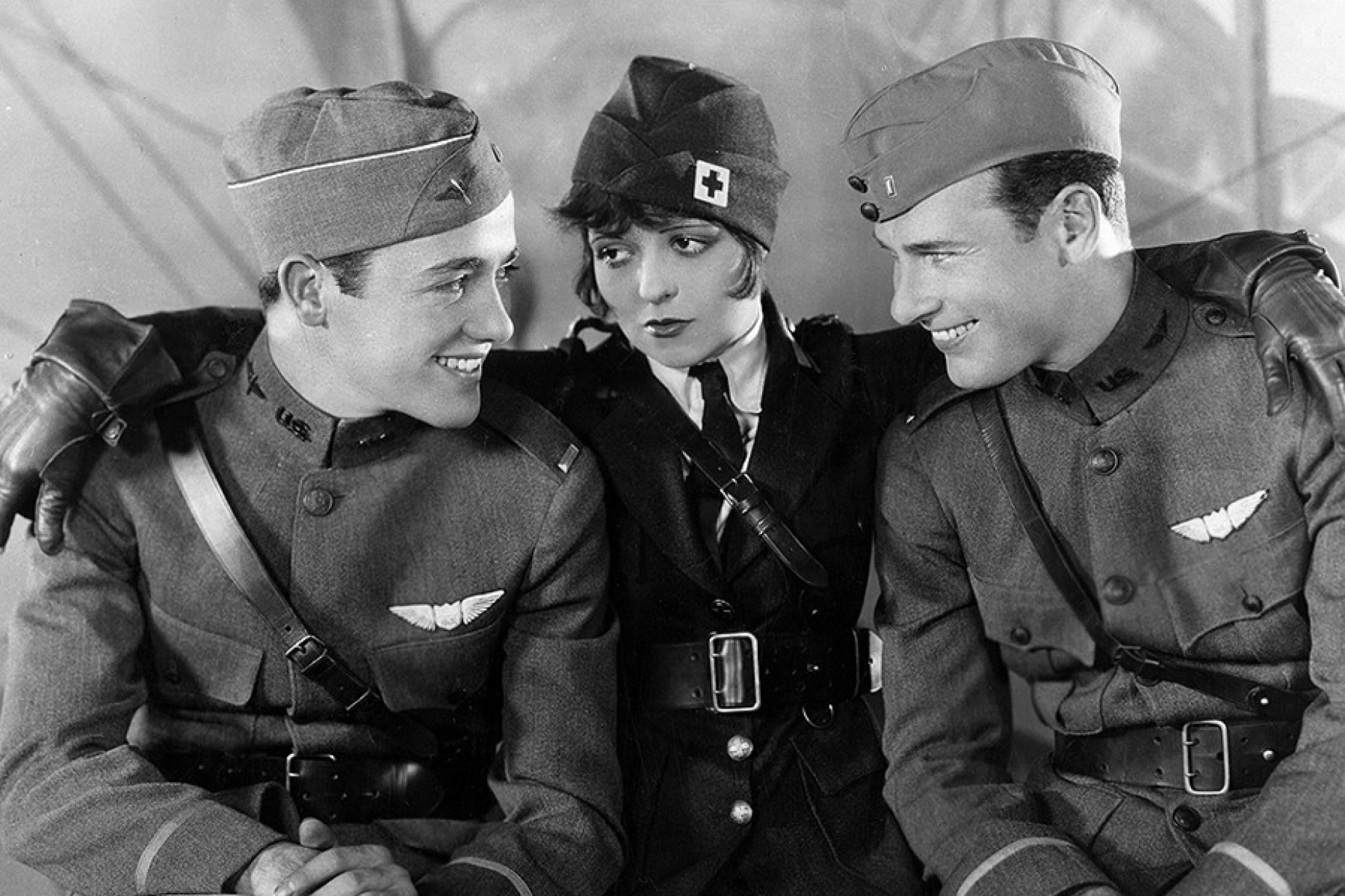 Still image from the 1927 movie, "Wings" featuring a woman with her arms around two men on either side, all wearing military uniforms. 