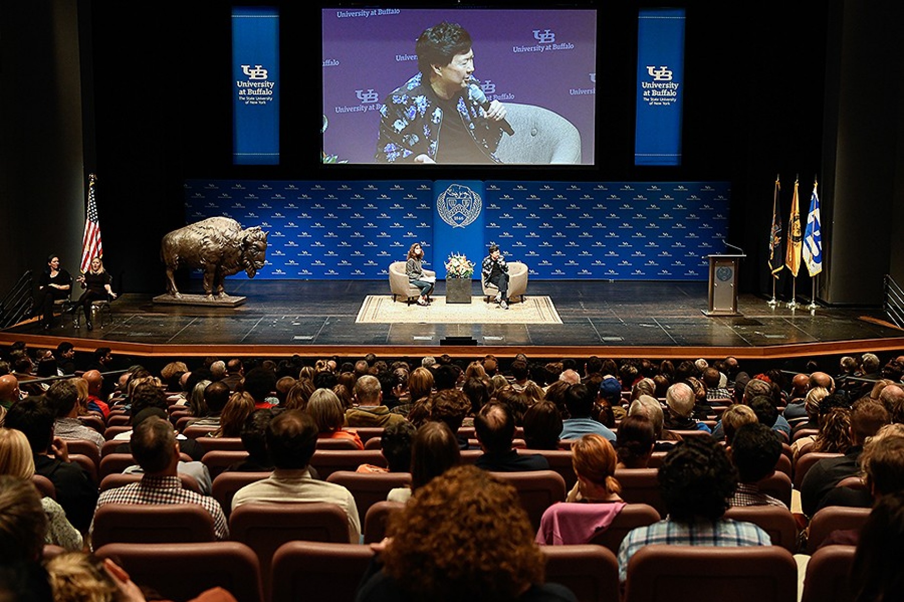 Ken Jeong, during his Distinguished speakers Series lecture, seated on stage and also projected on a large screen above. 