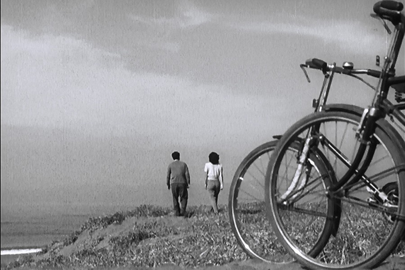 Scene from the movie, "Late Spring" featuring two bikes in the foreground as a couple walks towards a beach. 