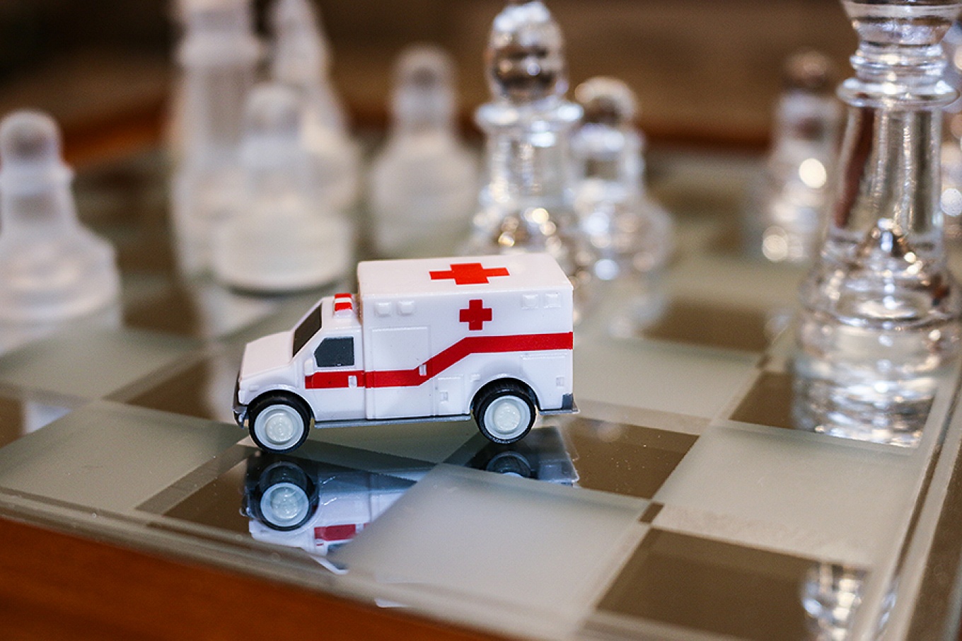 A white toy ambulance sits on a chessboard surrounded by glass chess pieces. 