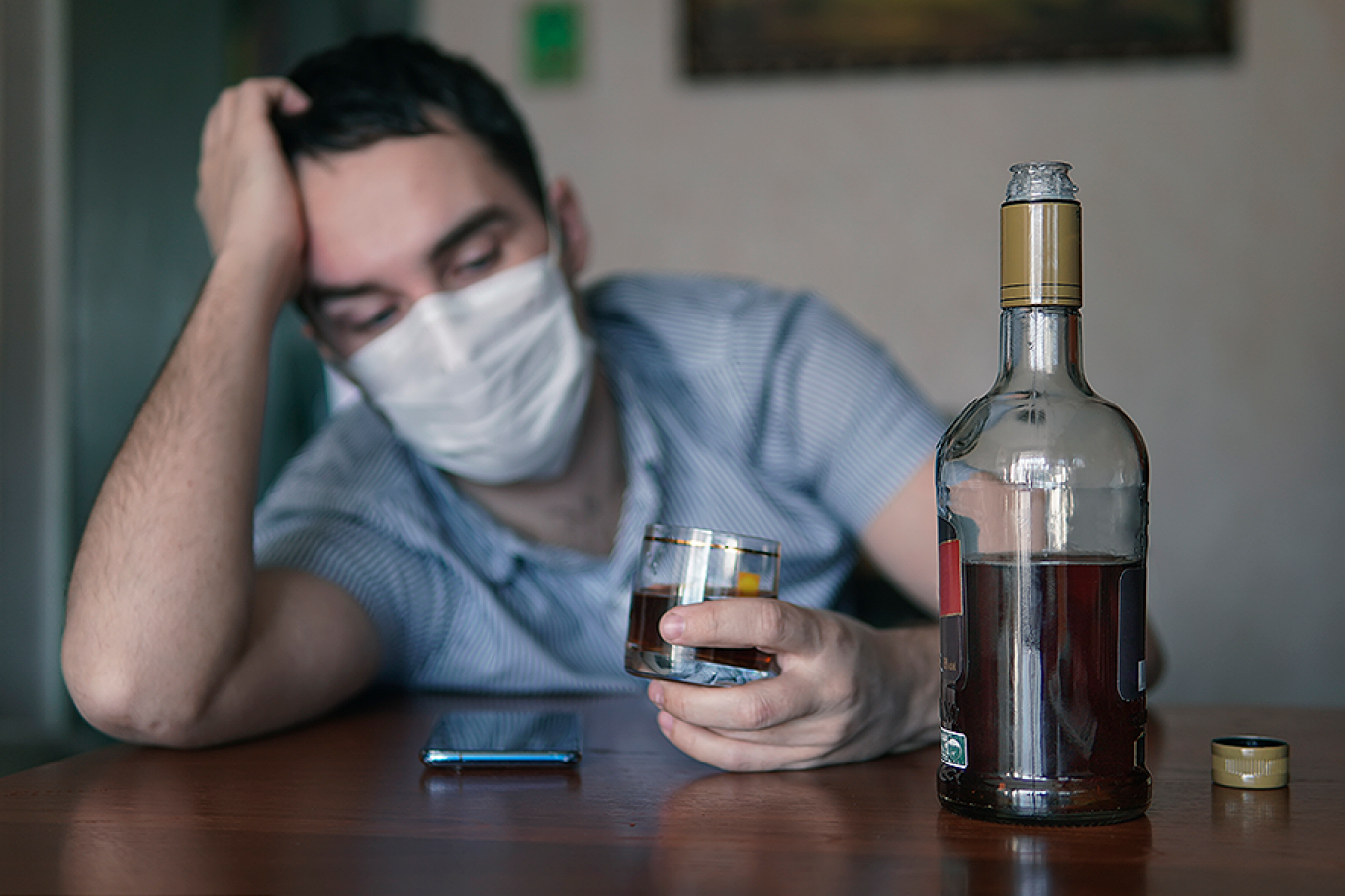 A depressed-looking person wearing a face mask holding a glass of liquor with a bottle of alcohol in the foreground. 