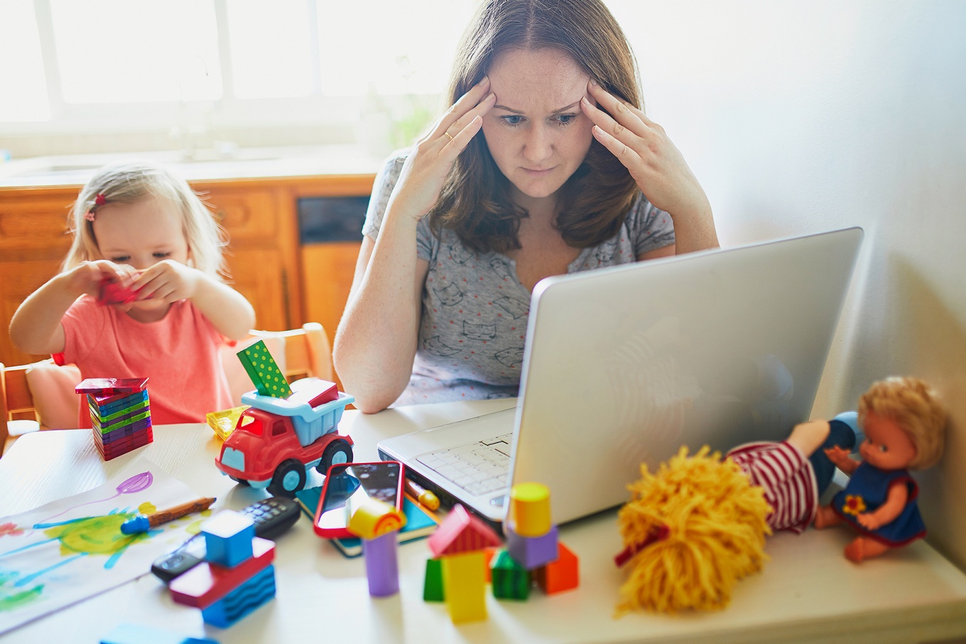 A young mother expresses frustration as she sits in front of a computer and her young child plays next to her. 