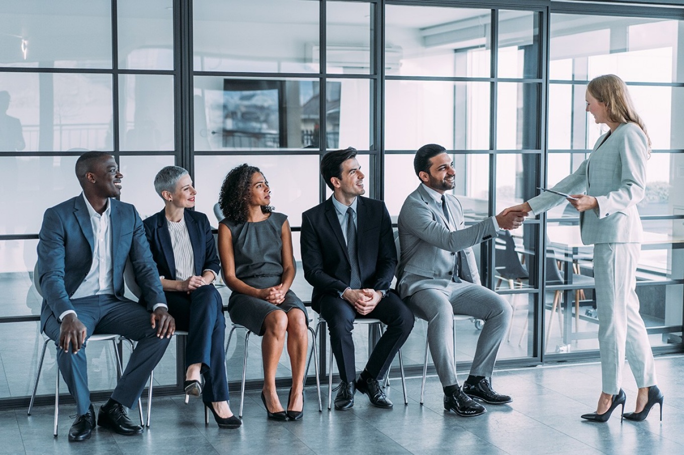 A diverse group of job candidates sit in a line as an interviewer shakes one person's hand.