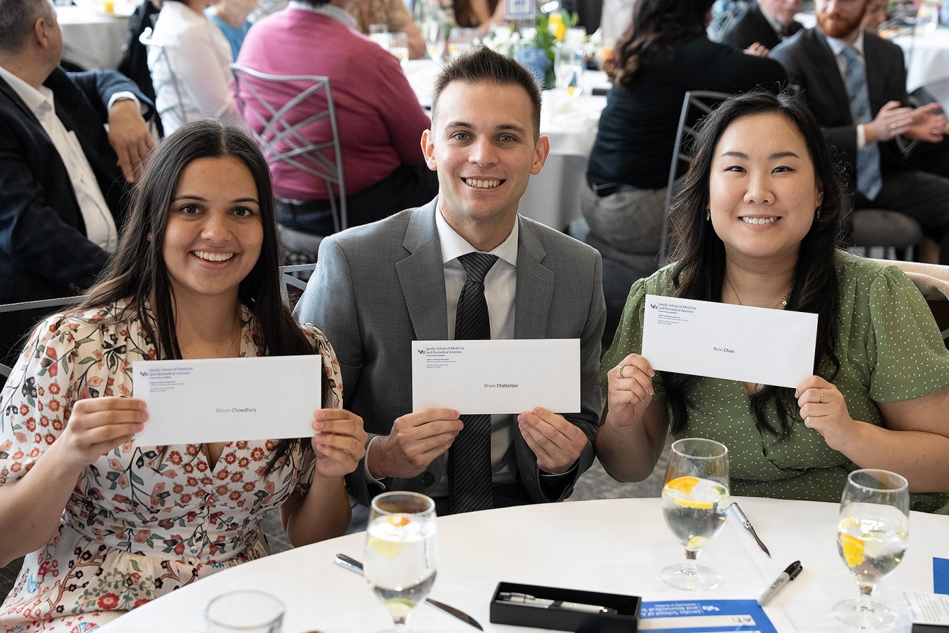 Three medical students, seated at a table, hold up their Match Day envelopes. 