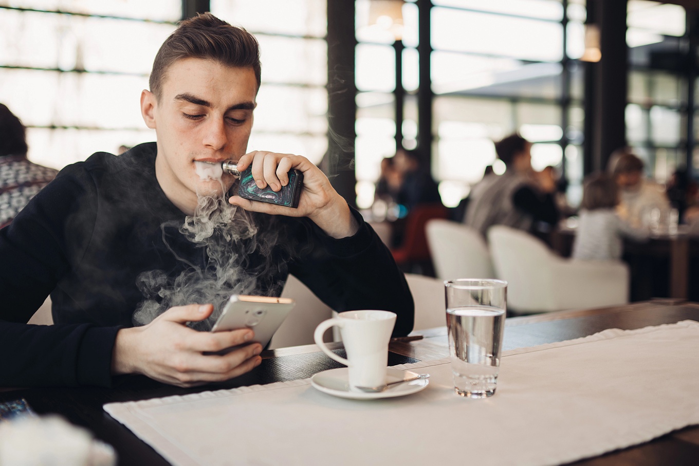 A young man vapes and looks at his phone while in a cafe. 