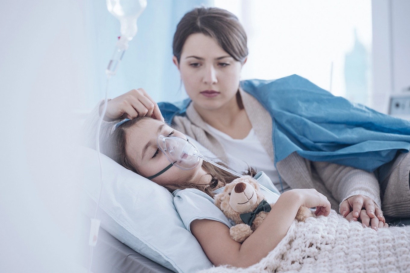 Mother lying next to a girl in bed wearing an oxygen mask. 