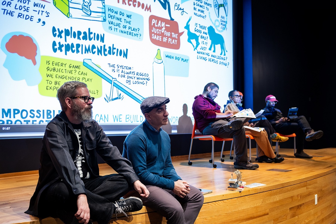 Twp people sit at the edge of a stage, behind there is a large projection screen and other people seated in chairs on stage. 