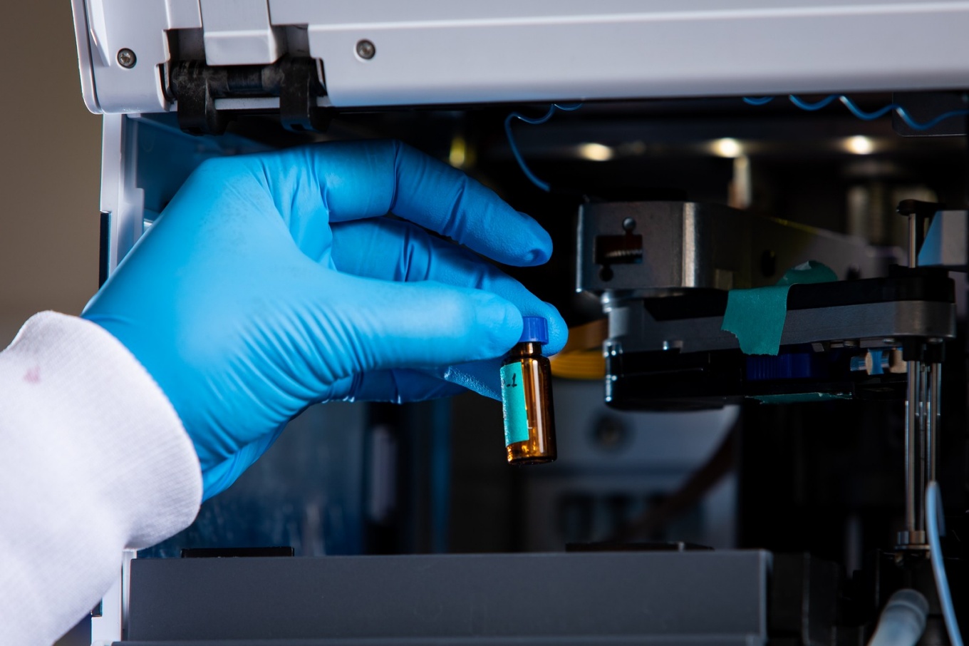Gloved hand holding a small vial near a piece of machinery.